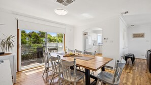 A modern kitchen with white cabinets, a dining area with a wooden table and chairs, and a sliding glass door leading to a balcony with greenery. - Falcon Bayview Beach House - Large Pet-Friendly Falcon Bayview Beach House with Ocean Views (Falcon)