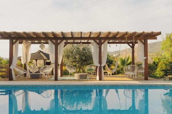 Ein Poolbereich mit einer Pergola, weißen Korbstühlen und Blick auf die Landschaft.