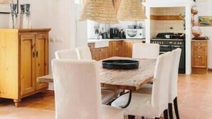 A dining area with a wooden table and white chairs, a kitchen in the background, and a hanging woven light fixture. - Oasis in tropical gardens with swimming pool (Moncarapacho)