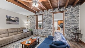Ein gemütliches Wohnzimmer mit Steinwand, Holzbalkendecke, einer Couch, einem Couchtisch und Blick in die Küche.