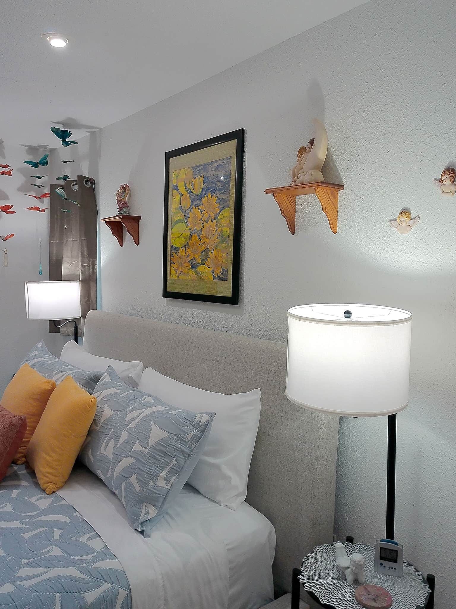 Quarto com cama, almofadas, abajur, prateleira na parede com objetos decorativos e um quadro emoldurado.