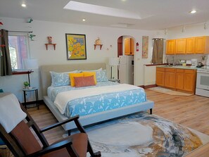 A bedroom with a bed, bedside tables, a chair, a small table, a window with curtains, a kitchen area with wooden cabinets, a refrigerator, and a stove. - Golden Harmony Cottage (Crestone)