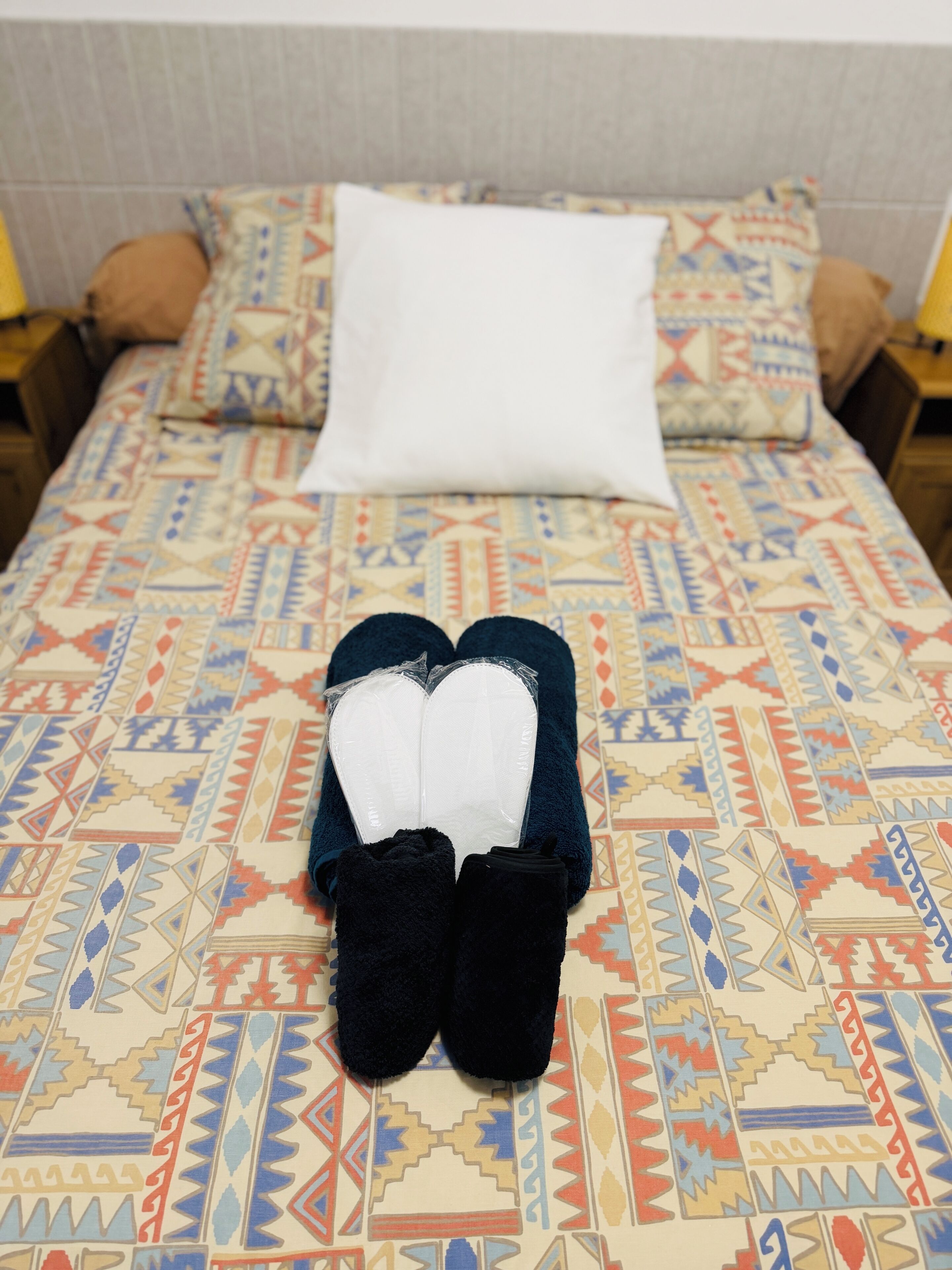 Een bed met een bedovertrek met dessin, een wit kussen en een paar zwarte pantoffels.
