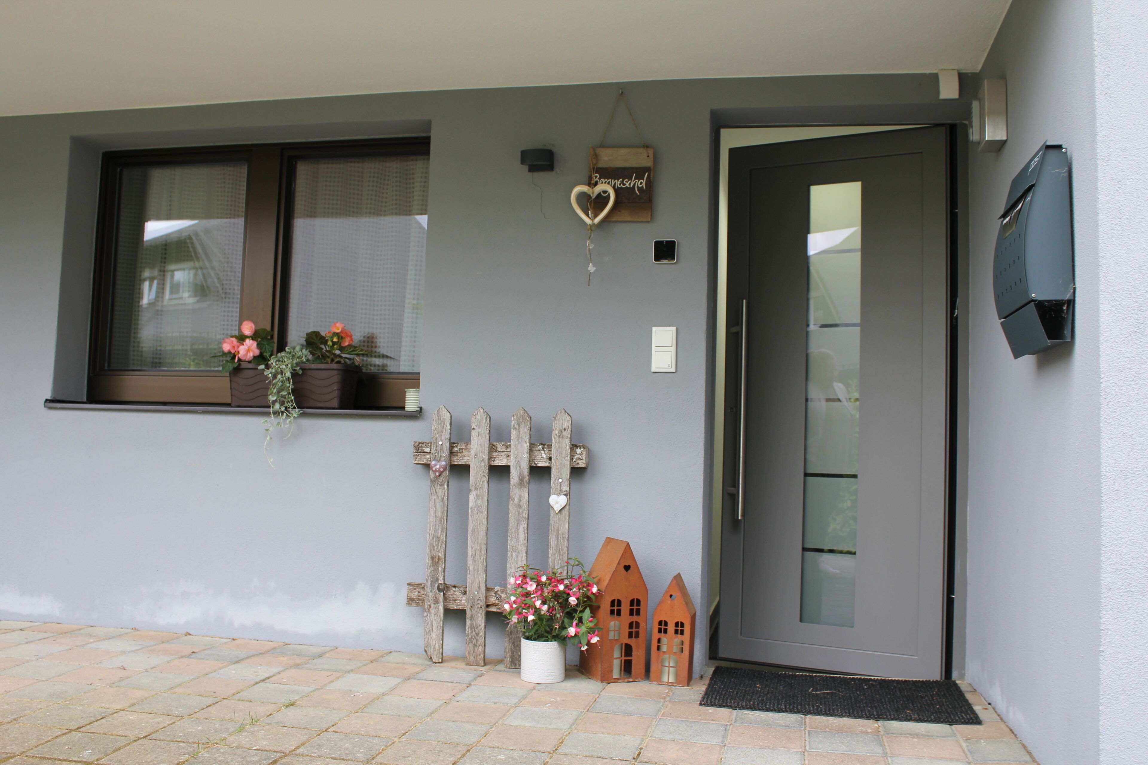 Pared exterior gris con una ventana, una puerta, un buzón pequeño y una planta en maceta.