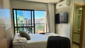 Ett hotellrum med en säng, en TV, en luftkonditioneringsanläggning, ett fönster med utsikt över byggnader och ett badrum som syns genom en öppen dörr.