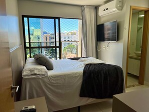 A hotel room with a bed, a TV, an air conditioning unit, a window with a view of buildings, and a bathroom visible through an open door. - Buenos Aires 504 - Apartment in the best location in Tambaú for 6 people. (João Pessoa)