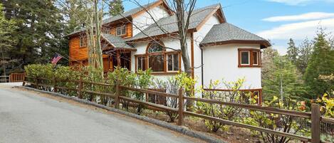 Une cabane en rondins à deux étages, avec une façade blanche, une terrasse en bois et une cour clôturée.