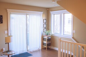 Une pièce avec un plancher en bois, un bureau avec une lampe, une chaise, une fenêtre avec des rideaux et une plante posée sur une étagère.