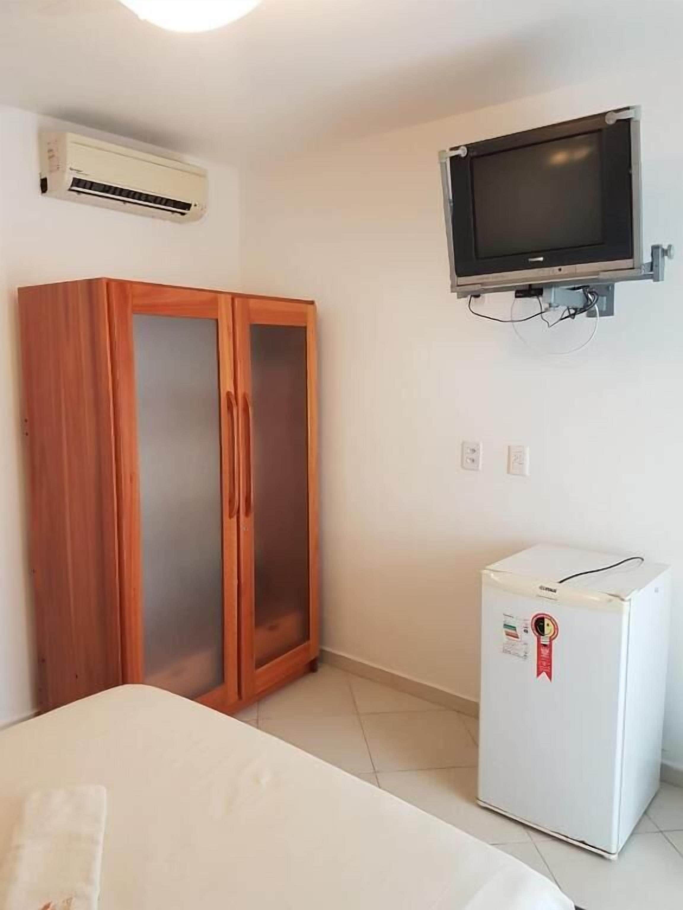 A room with a wooden wardrobe, a white bed, a small white refrigerator, and a mounted television.