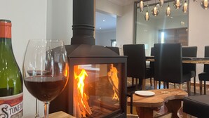A wood-burning stove with visible flames, a glass wine bottle, and a wine glass on a wooden table in a dining area with chairs and a large mirror.