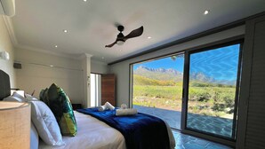 A bedroom with a bed, a ceiling fan, a large window with a mountain view, and a stone floor.