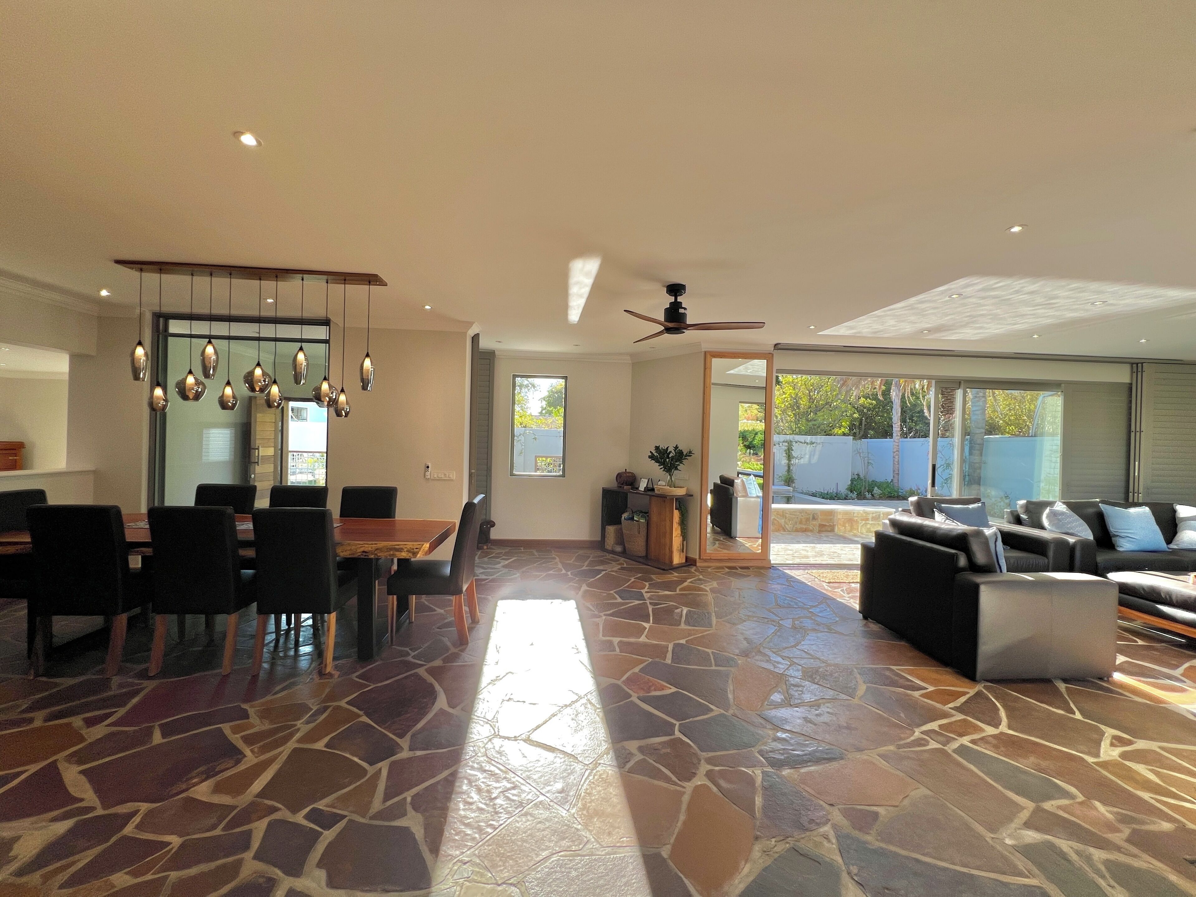 A spacious living area with a dining table, black chairs, a stone-tiled floor, a ceiling fan, and a large sliding glass door leading to a patio.
