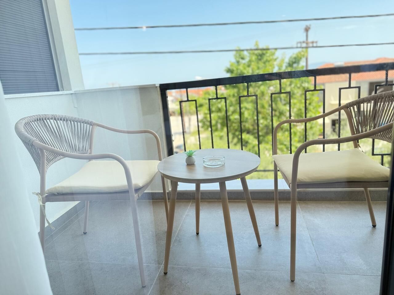 Een balkon met twee rieten stoelen, een kleine ronde tafel, een bloempot en uitzicht op groen en gebouwen.