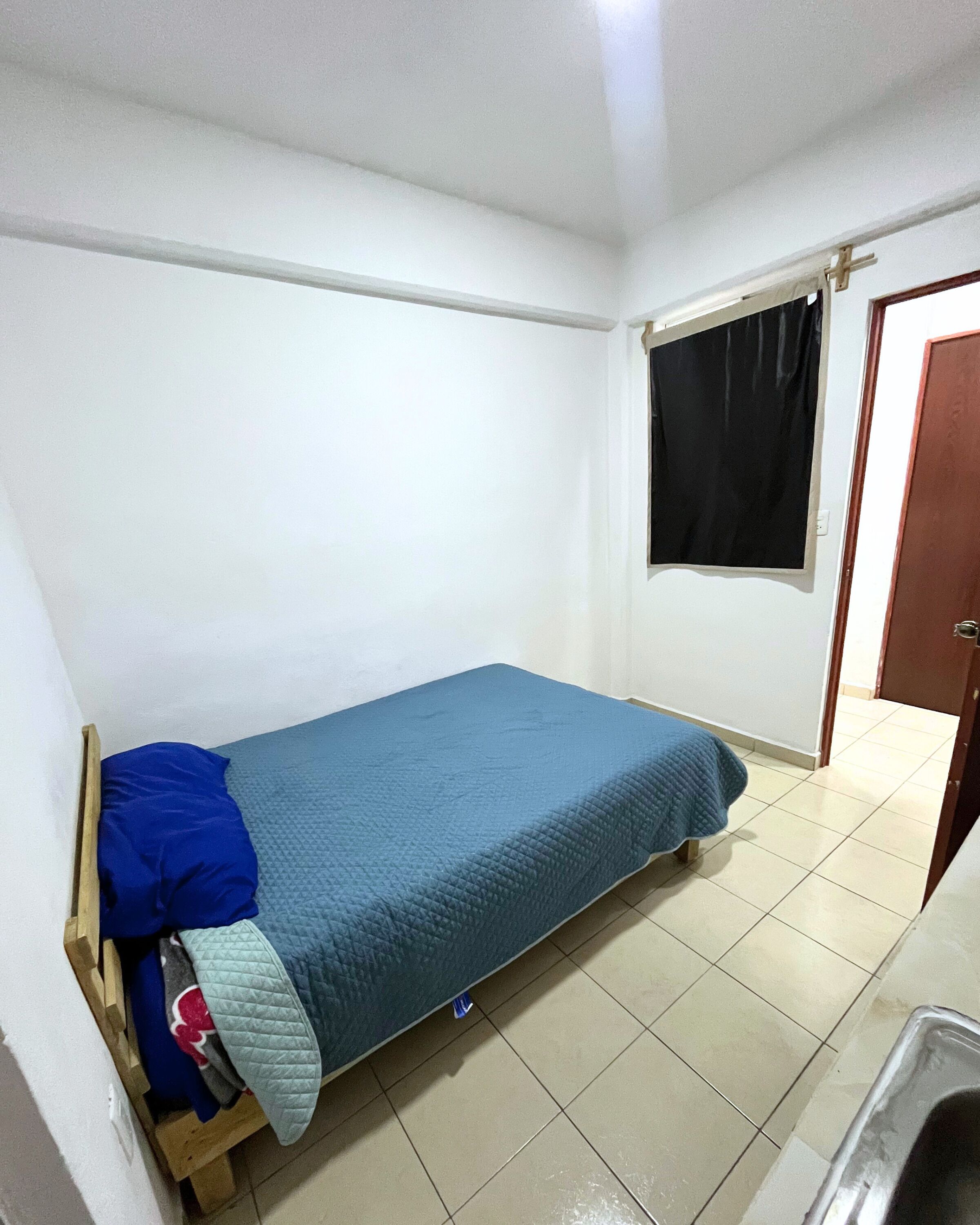 A small, single-bed room with a wooden frame, a blue mattress, and a black curtain on the window.