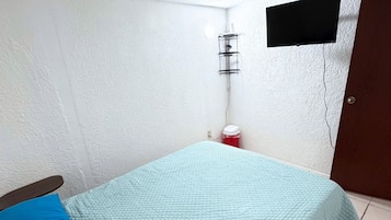 Une pièce avec un lit, un oreiller bleu, une poubelle rouge, un téléviseur noir fixé au mur et une porte en bois.