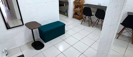 Una stanza con una panca verde acqua, un tavolino e una sedia nera. Si vede una parete bianca con uno specchio e un armadietto di legno.