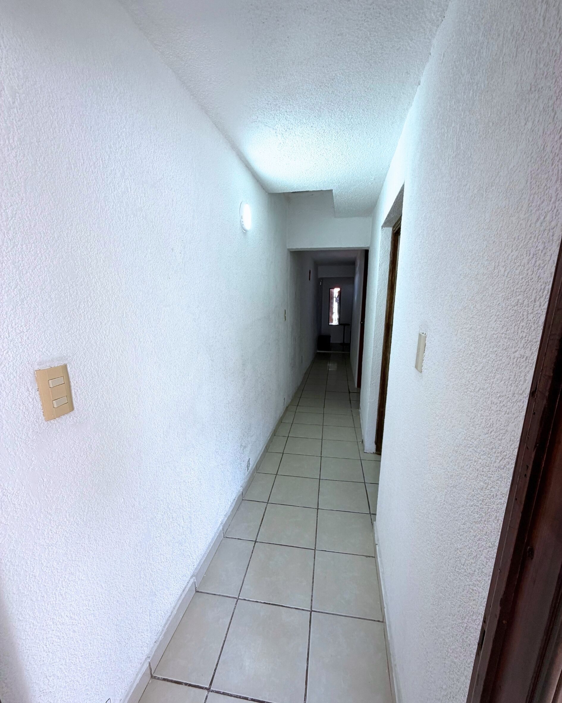 Un pasillo estrecho con paredes blancas y piso de baldosas que conduce a una puerta al final.