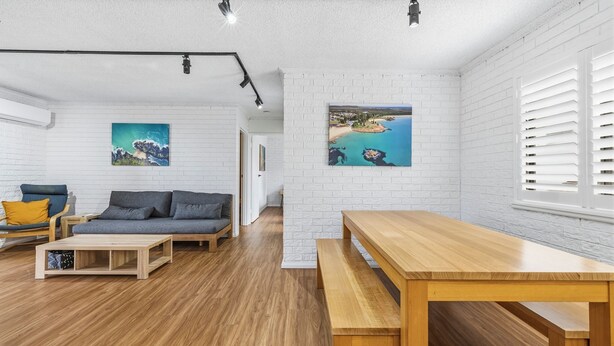 Ein modernes Wohnzimmer mit einer Couch, einem Couchtisch und einer Holzbank. Weiße Ziegelwände und ein großes Fenster mit Jalousien.