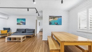 A modern living room with a sofa, coffee table, and wooden bench. There are white brick walls and a large window with blinds. - Azure - A comfortable holiday unit designed for rest, relaxation, and convenience (South West Rocks)