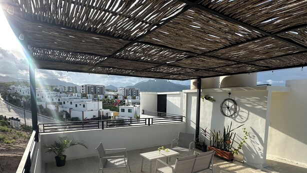 Een balkon met een pergola, zitplaatsen, een tafel, potplanten en een klok aan de muur.