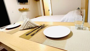 Een hotelkamer met een netjes opgemaakte eettafel, een bed met een witte dekbedovertrek, een aan de muur bevestigde televisie en een houten plankje met een klein voorwerp erop.