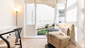 Ein Zimmer mit einer Couch, einem kleinen Tisch, einer Lampe und Blick auf eine weiße Backsteinmauer und einen Garten.