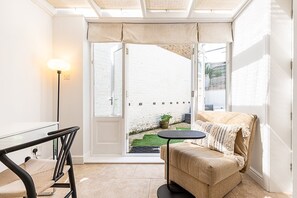 A room with a sofa, a small table, a lamp, and a view of a white brick wall and garden outside. - Stylish Fulham Apartment with Private Garden (London)
