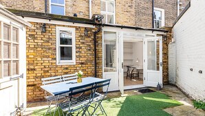 Ein Ziegelhaus mit weißer Tür, eine kleine Terrasse mit Tisch und Stühlen sowie ein Rasengrundstück.