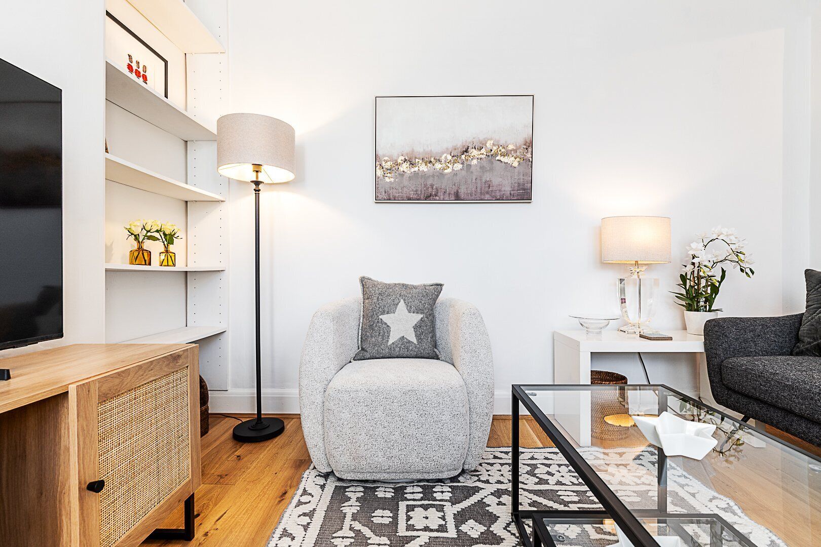 Een moderne woonkamer met een grijze bank, een kussen met stermotief, een vloerlamp, een lijstwerk en een televisie op een houten kast.