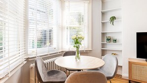 Ein modernes Zimmer mit einem runden Tisch, grauen Stühlen, einem Heizkörper, weißen Regalen und einem Fernseher.