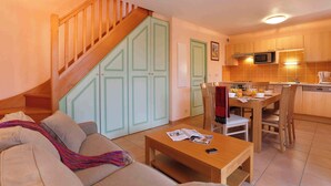 Una sala de estar con un sofá, una mesa de centro y un comedor con una mesa y sillas. Una escalera con barandales de madera lleva a una puerta verde.