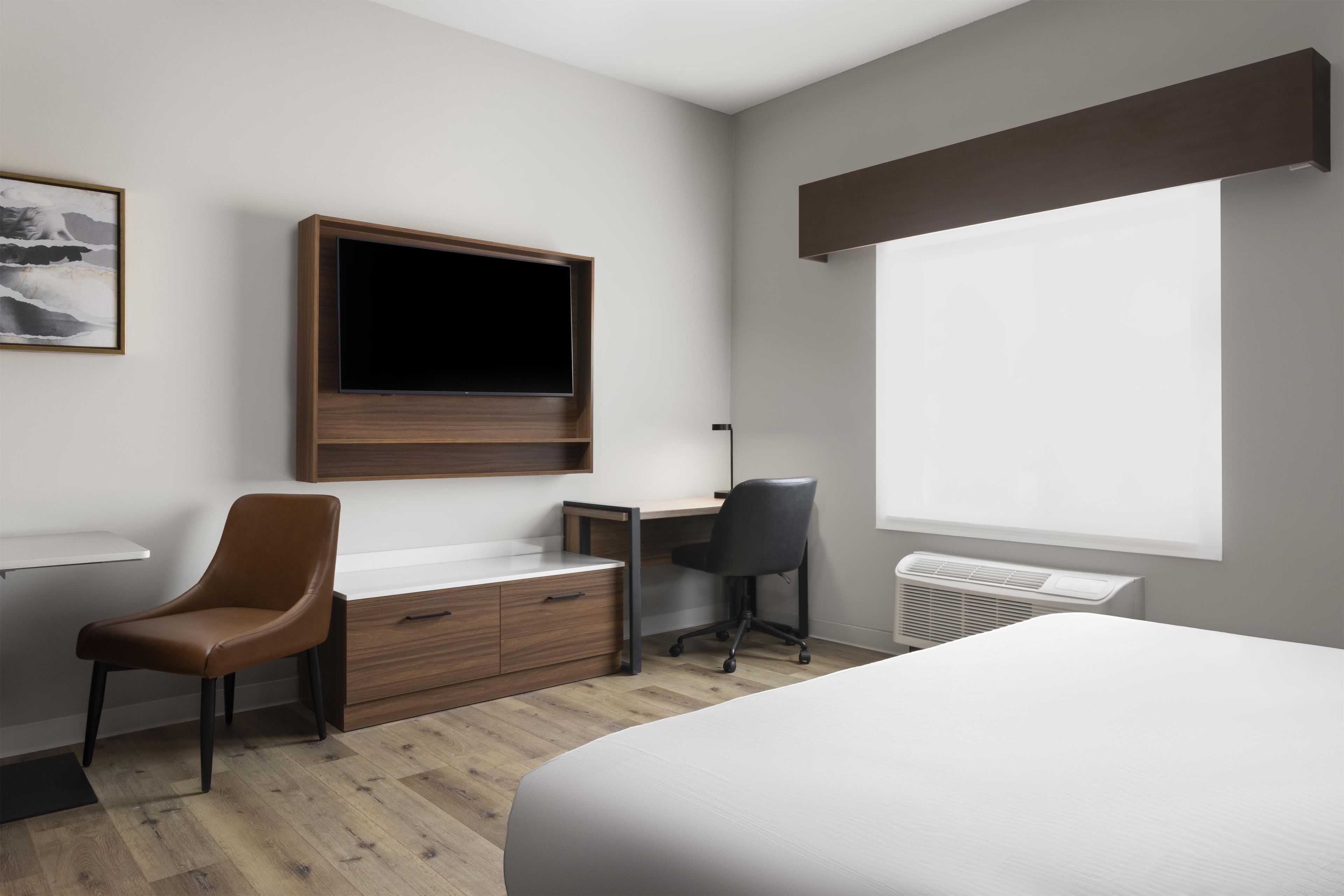 A hotel room with a bed, a desk with a chair, a television mounted on the wall, and a window.