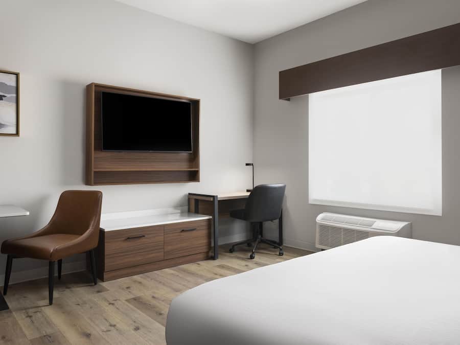 Quarto de hotel com cama, escrivaninha e cadeira, televisão na parede e janela.