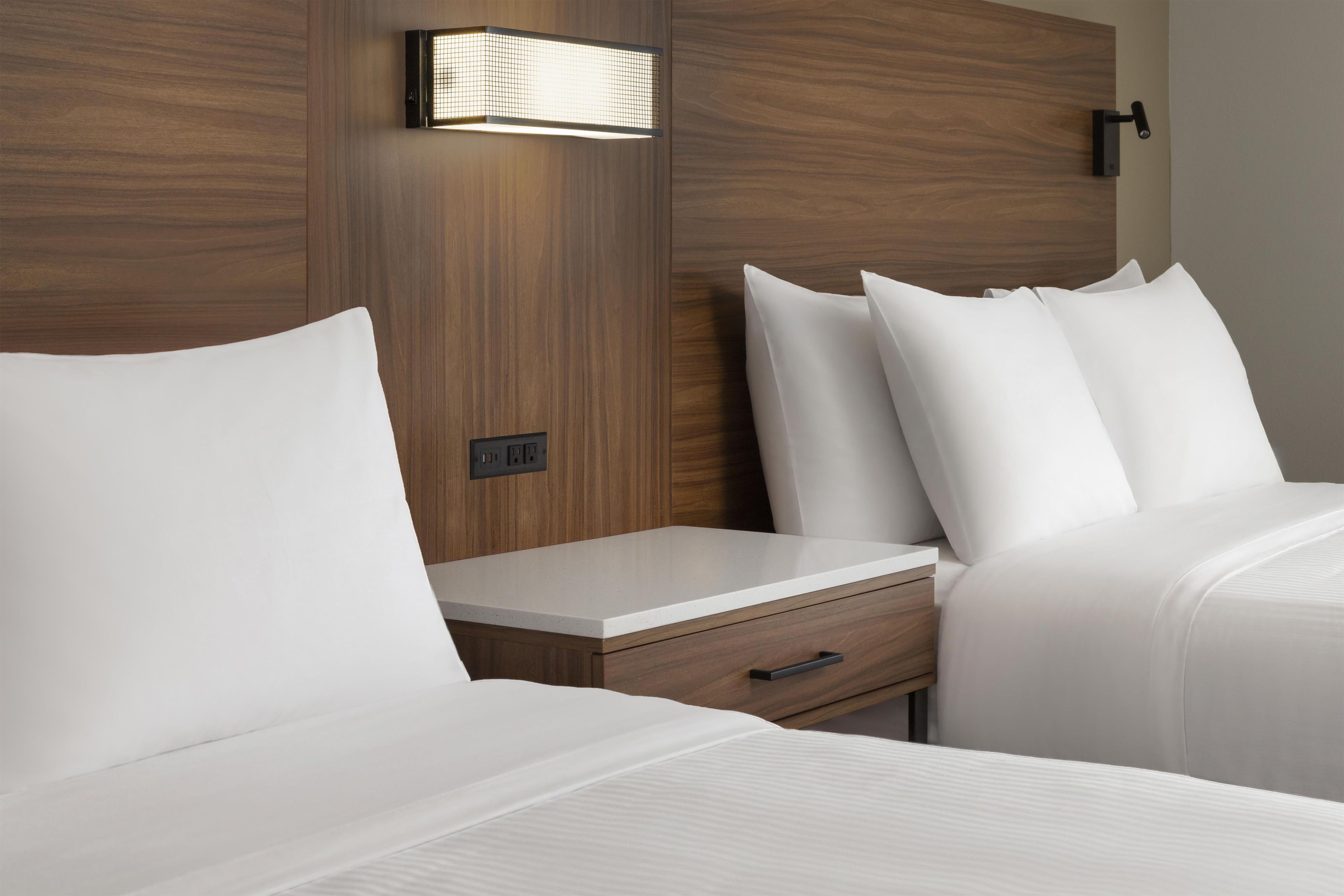 A hotel room with a wooden headboard, a bedside table with a drawer, and a wall-mounted light fixture.