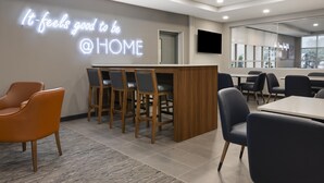 A modern interior with a neon sign reading 'It feels good to be @ HOME', a reception desk, and a dining area with chairs and tables.