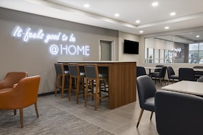 A modern interior with a neon sign reading 'It feels good to be @ HOME', a reception desk, and a dining area with chairs and tables.