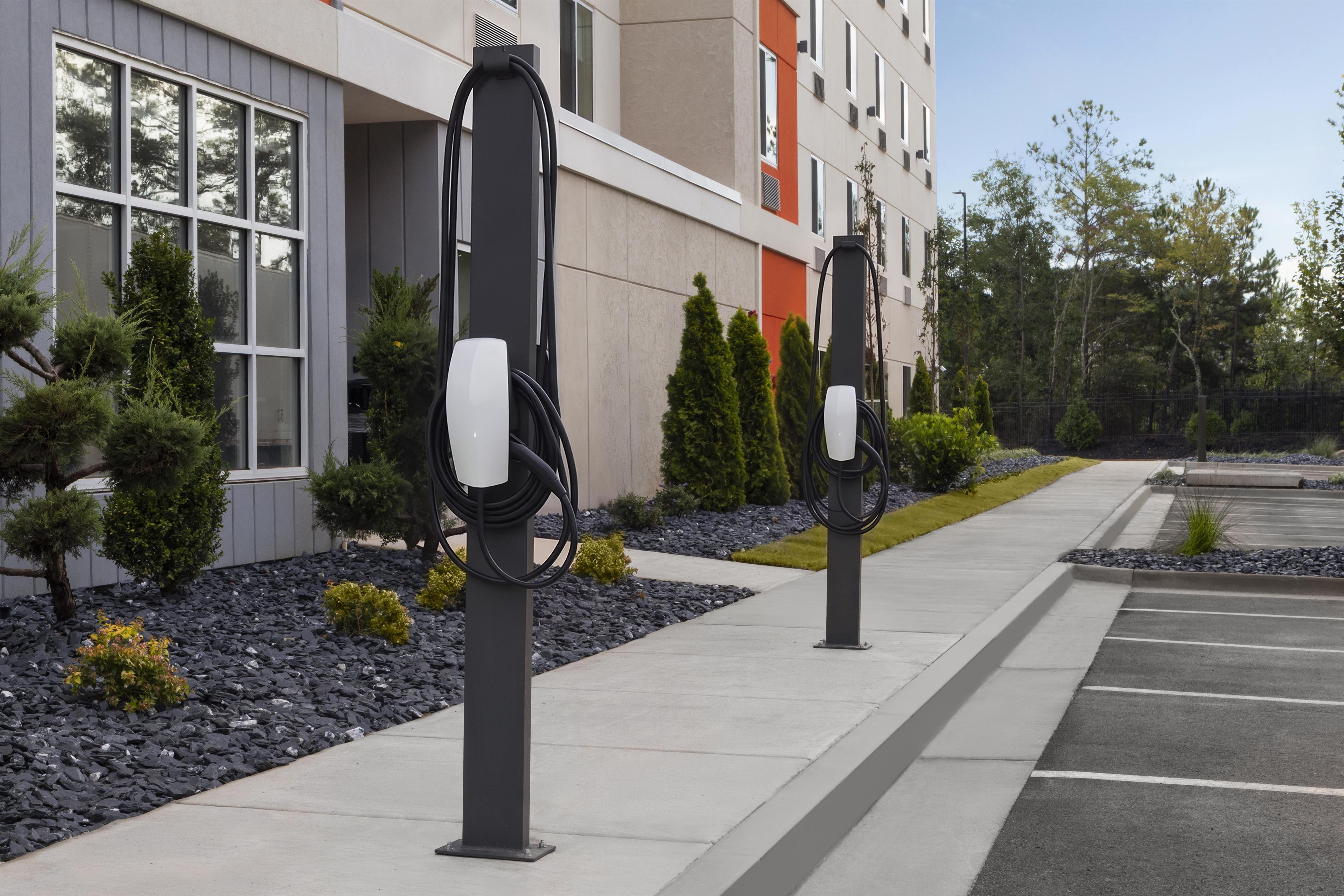 Dos estaciones de carga para vehículos eléctricos en una acera, con un edificio y árboles al fondo.