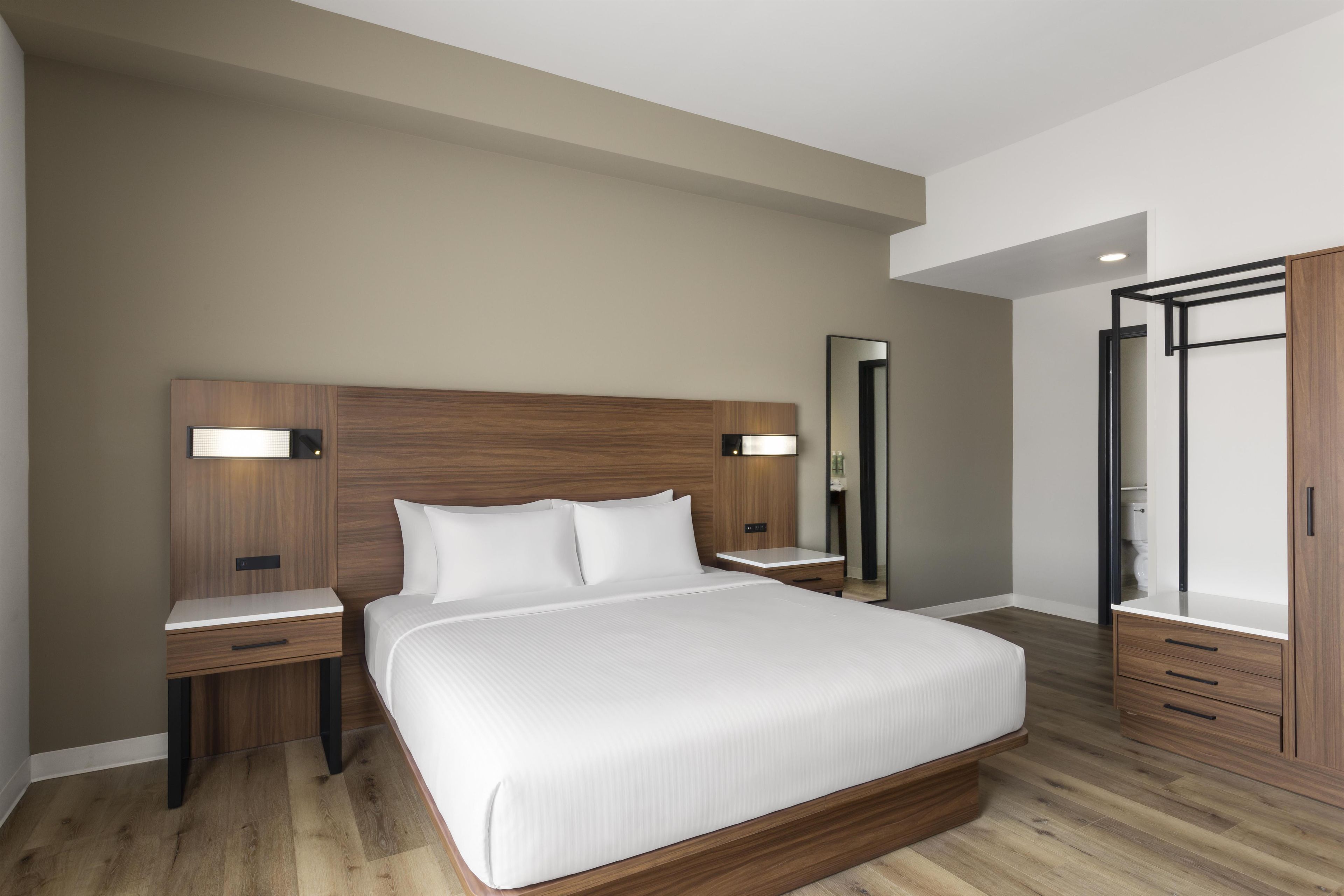 A modern hotel room with a large bed, wooden headboard, bedside tables, a mirror, and a wardrobe.