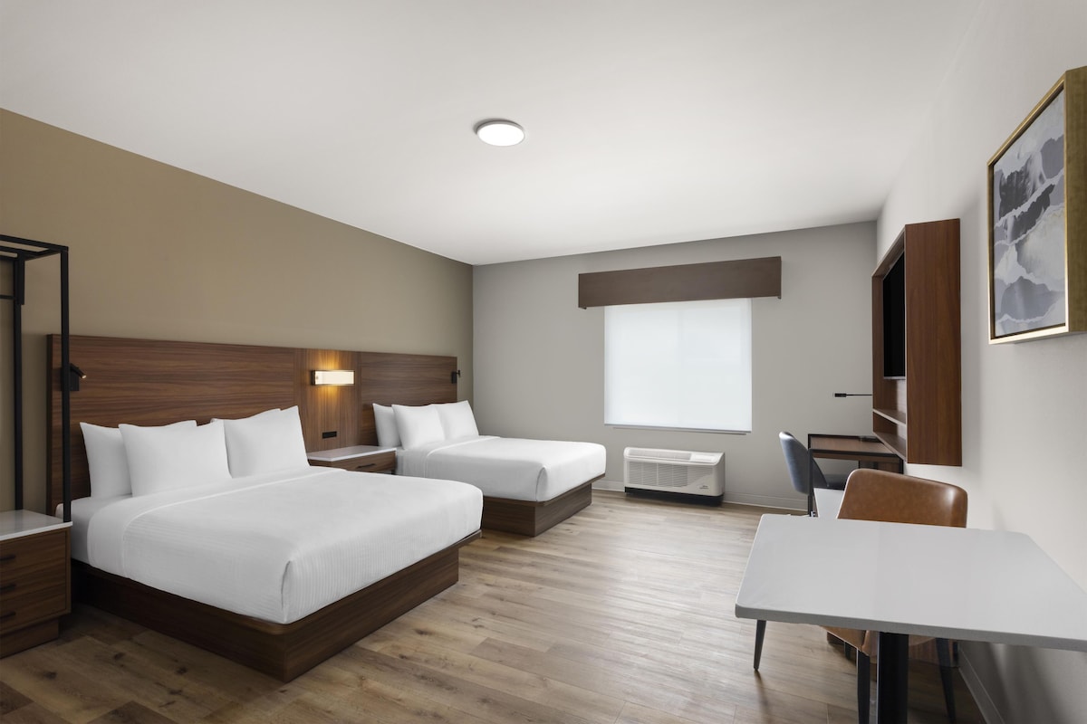 A hotel room with two beds, a desk, a chair, a window, and a mounted artwork.