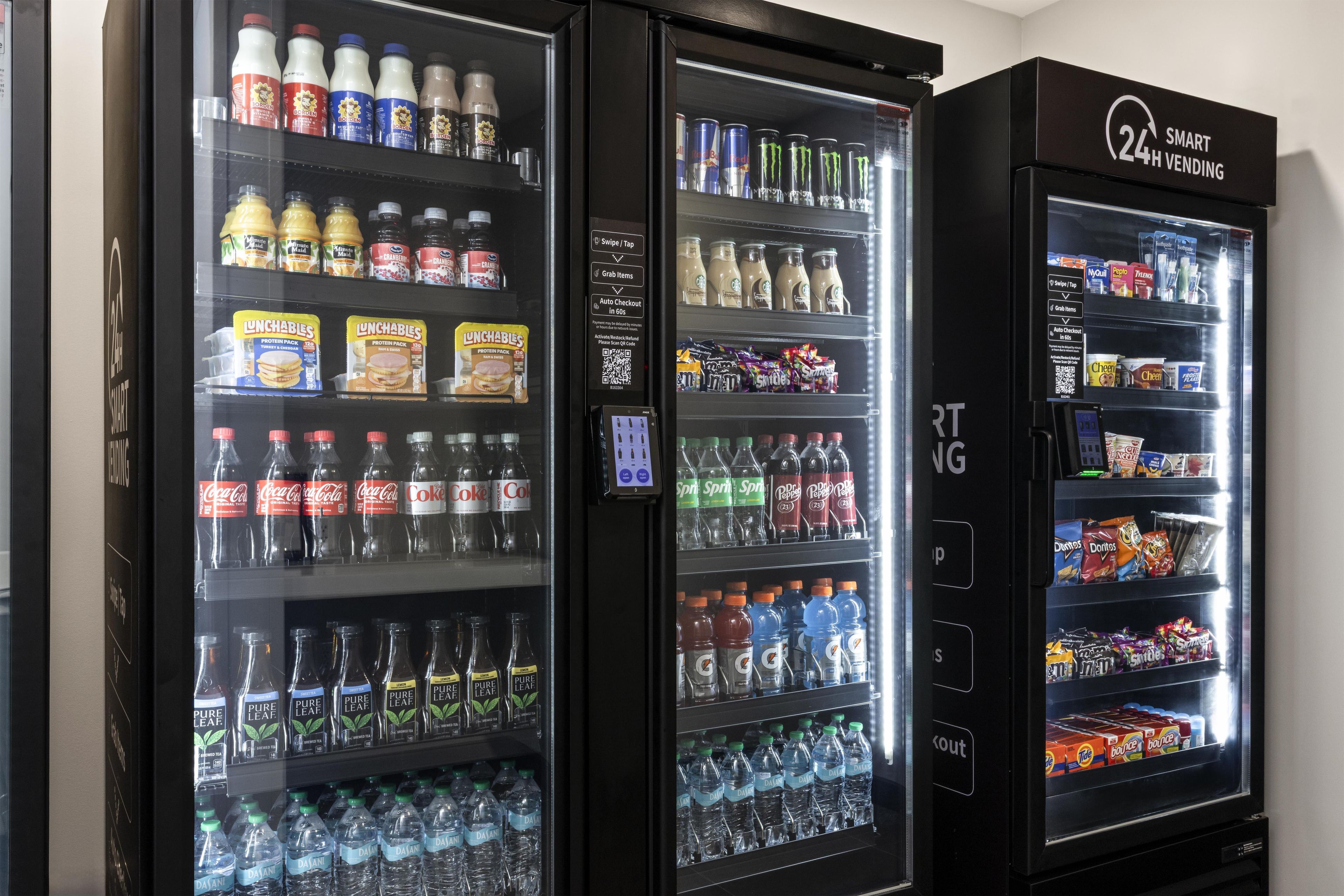 Trois distributeurs automatiques de boissons proposent une variété de produits, notamment des sodas, des jus de fruits et de l’eau en bouteille.