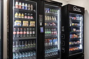 Trois distributeurs automatiques de boissons proposent une variété de produits, notamment des sodas, des jus de fruits et de l’eau en bouteille.