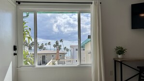 Ein Zimmer mit großem Fenster, Blick auf Palmen und Gebäude, einem kleinen Tisch mit einer Pflanze und einem an der Wand montierten Fernseher.