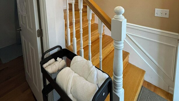 Eine Treppe mit Holzdielen und weißen Spindeln, ein schwarzer Metallstuhl mit weißem Sitzkissen und eine Tür, die in einen anderen Raum führt.