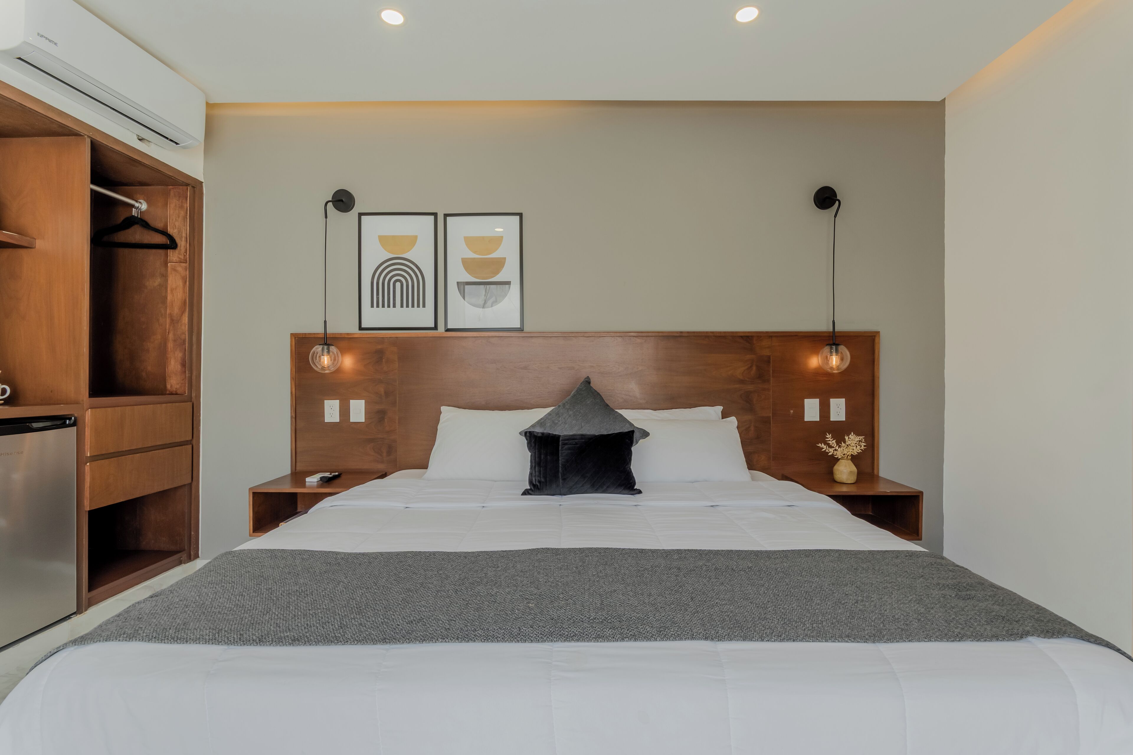A hotel room with a bed, wooden wardrobe, bedside table, and wall-mounted artwork.