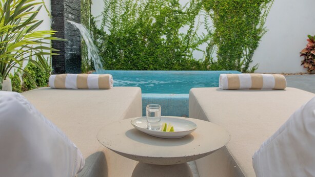 A poolside lounge with a table, cushions, a glass of water, and a plate of fruit.