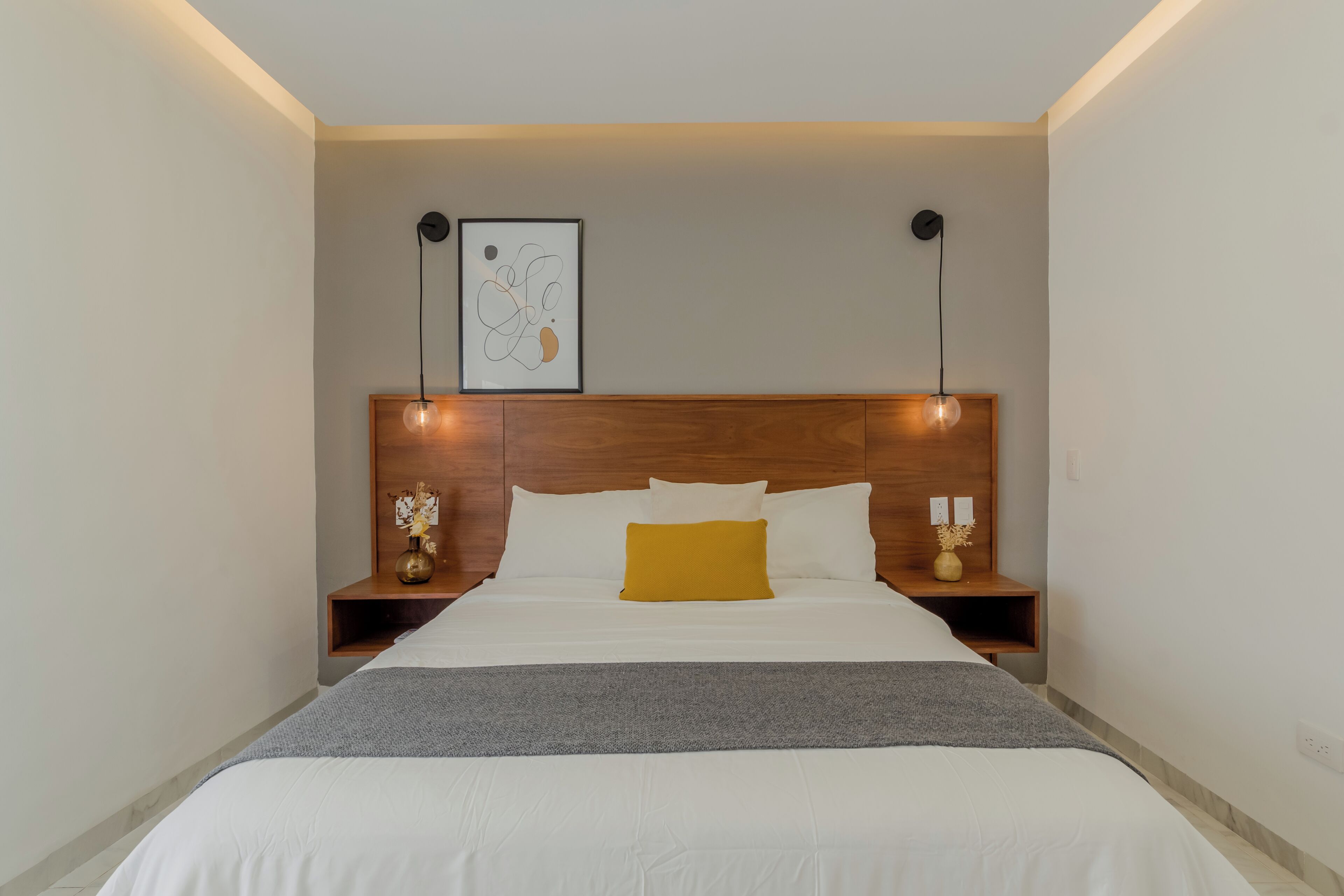 A neatly made bed with a white and yellow pillow, a gray throw blanket, and a wooden headboard. There are two bedside tables with lamps and a framed abstract art piece above the bed.