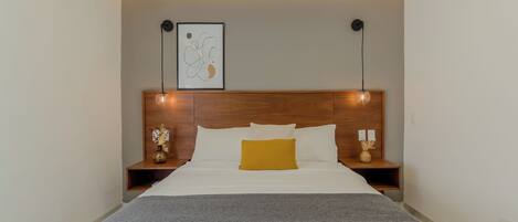 A neatly made bed with a white and yellow pillow, a gray throw blanket, and a wooden headboard. There are two bedside tables with lamps and a framed abstract art piece above the bed.