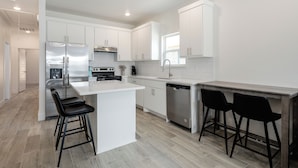 A modern kitchen with white cabinetry, a stainless steel refrigerator, a dishwasher, and two black bar stools. - Brand New Home Near Downtown (Sulphur Springs)