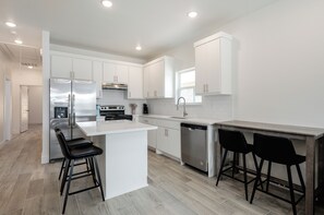 A modern kitchen with white cabinetry, a stainless steel refrigerator, a dishwasher, and two black bar stools. - Brand New Home Near Downtown (Sulphur Springs)