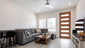 A modern living room with a grey sectional sofa, a wooden coffee table, a black bar stool, a wooden door, and a television on a stand. - Brand New Home Near Downtown (Sulphur Springs)